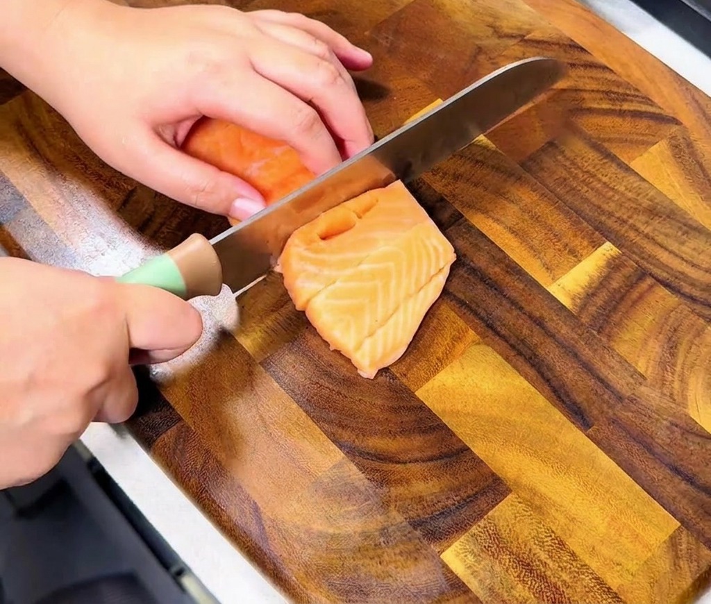 Hände schneiden ein frisches Lachsfilet auf einem Holzbrett in gleichmäßige, mundgerechte Würfel.