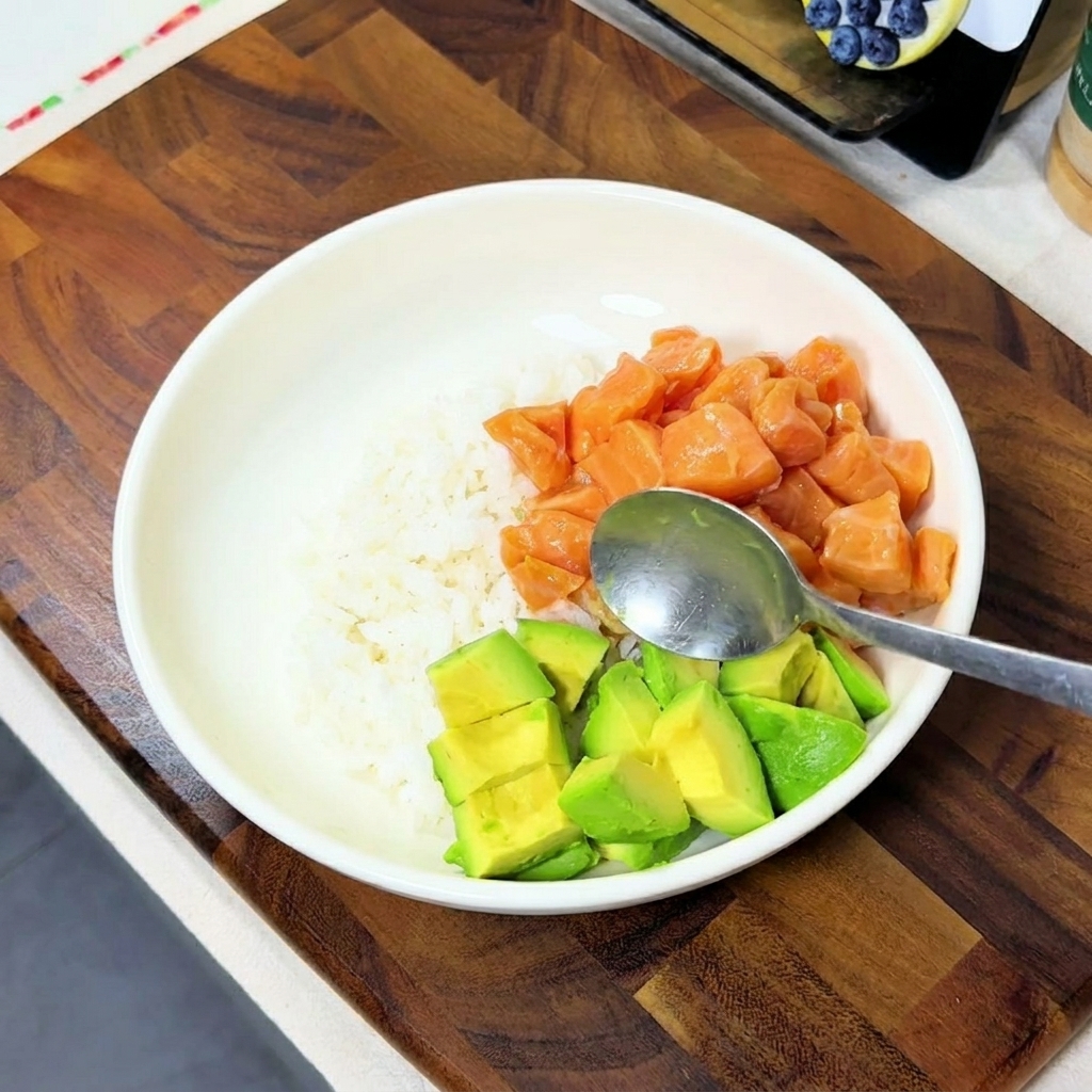 Avocadowürfel werden mit einem Löffel in die Bowl zu Reis und Lachs gegeben.