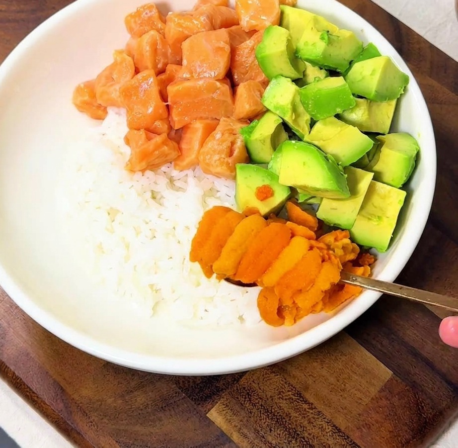 Eine Hand platziert orangefarbene Seeigelstücke auf die Bowl mit Lachs und Avocado.