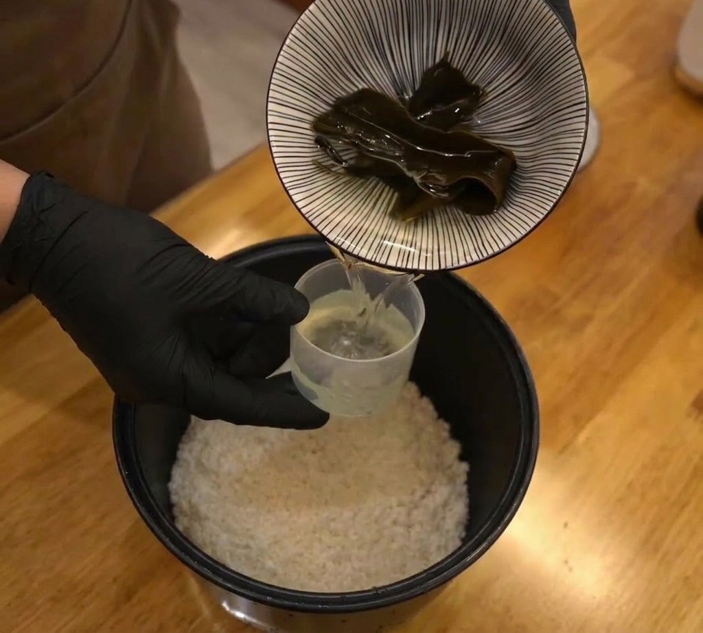 Ein Stück dunkler, getrockneter Kombu wird in den Reiskocher zu trockenem Reis und Wasser gegeben.