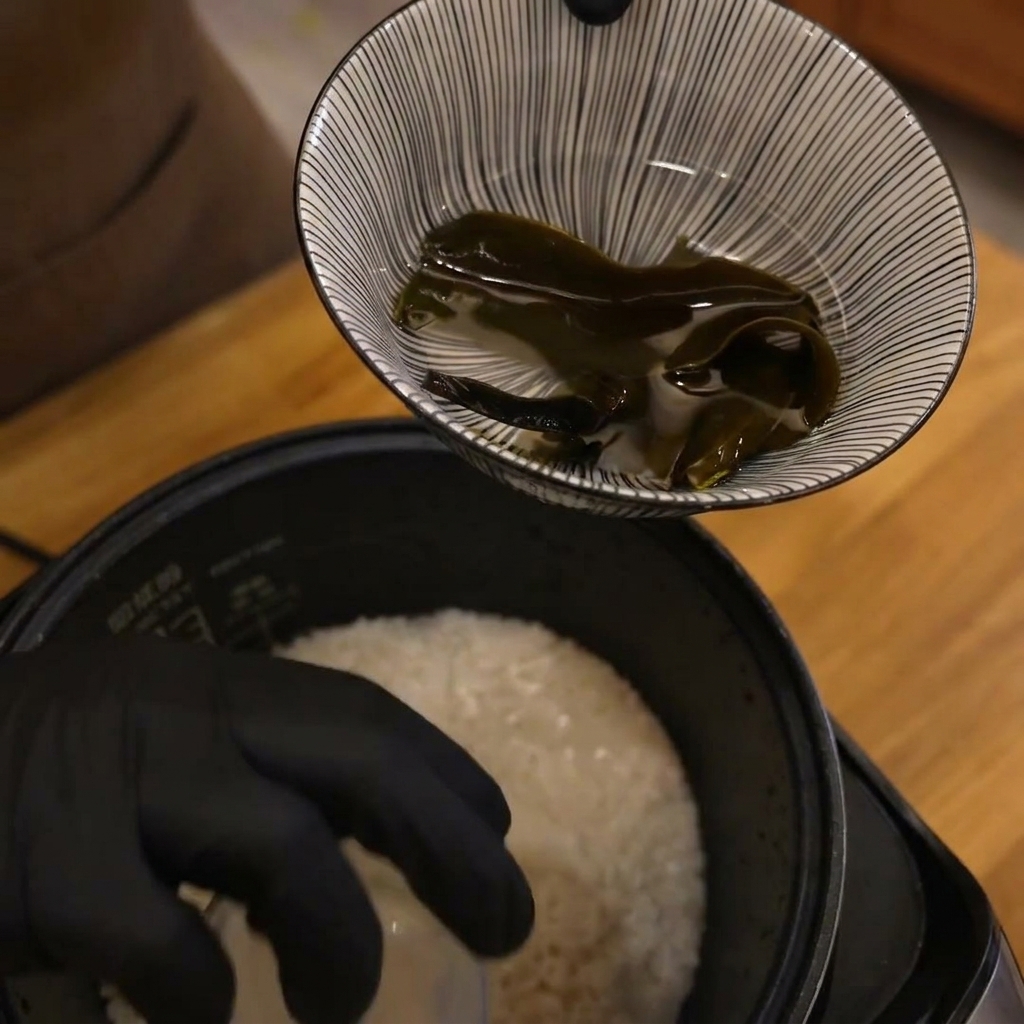 Eine behandschuhte Hand hält einen Messbecher über dem Reiskocher, um Wasser zum Reis hinzuzufügen.