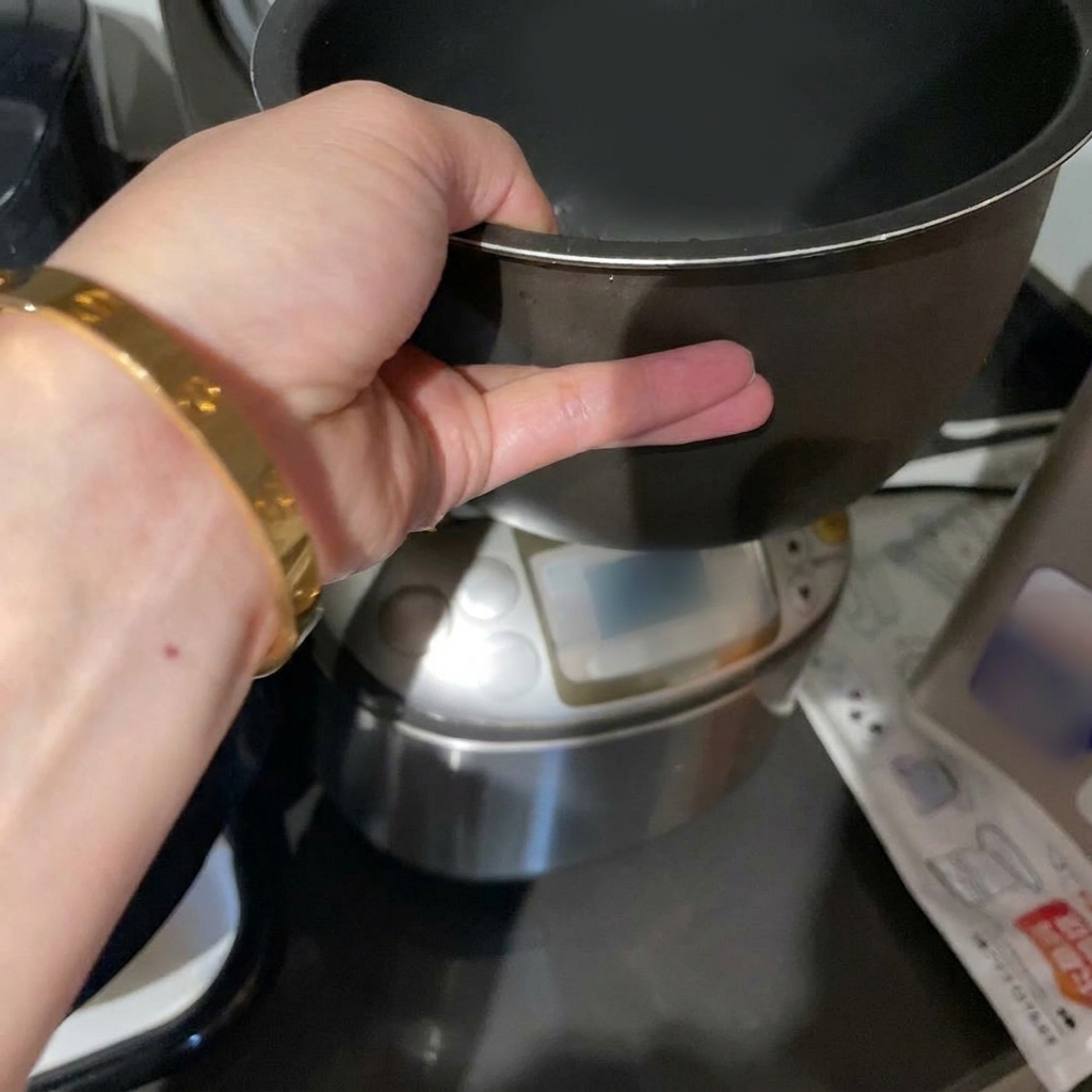 Eine Hand hält den dunklen Innentopf eines Reiskochers mit gewaschenem weißem Reis unter Wasser.