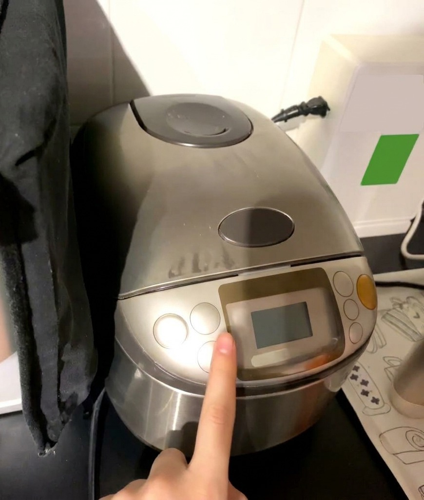Ein Finger drückt die Starttaste an einem silbernen Reiskocher mit digitalem Display.