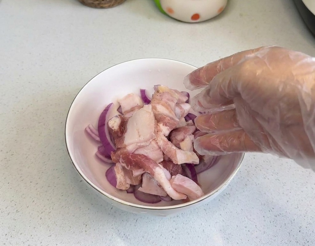 Eine Hand mit einem transparenten Plastikhandschuh legt rohe Schweinebauchscheiben in eine weiße Schüssel mit roten Zwiebelringen.