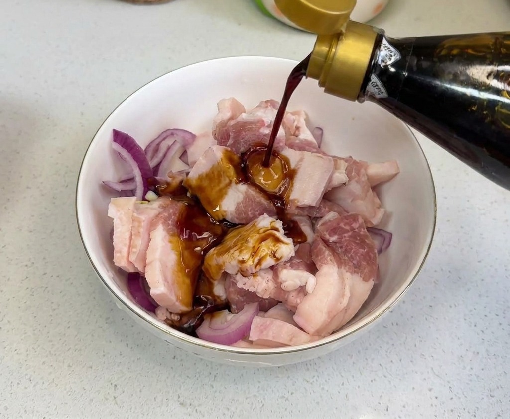 Dunkle Sojasauce wird aus einer Glasflasche über rohes Fleisch und geschnittene Zwiebeln in einer Rührschüssel gegossen.