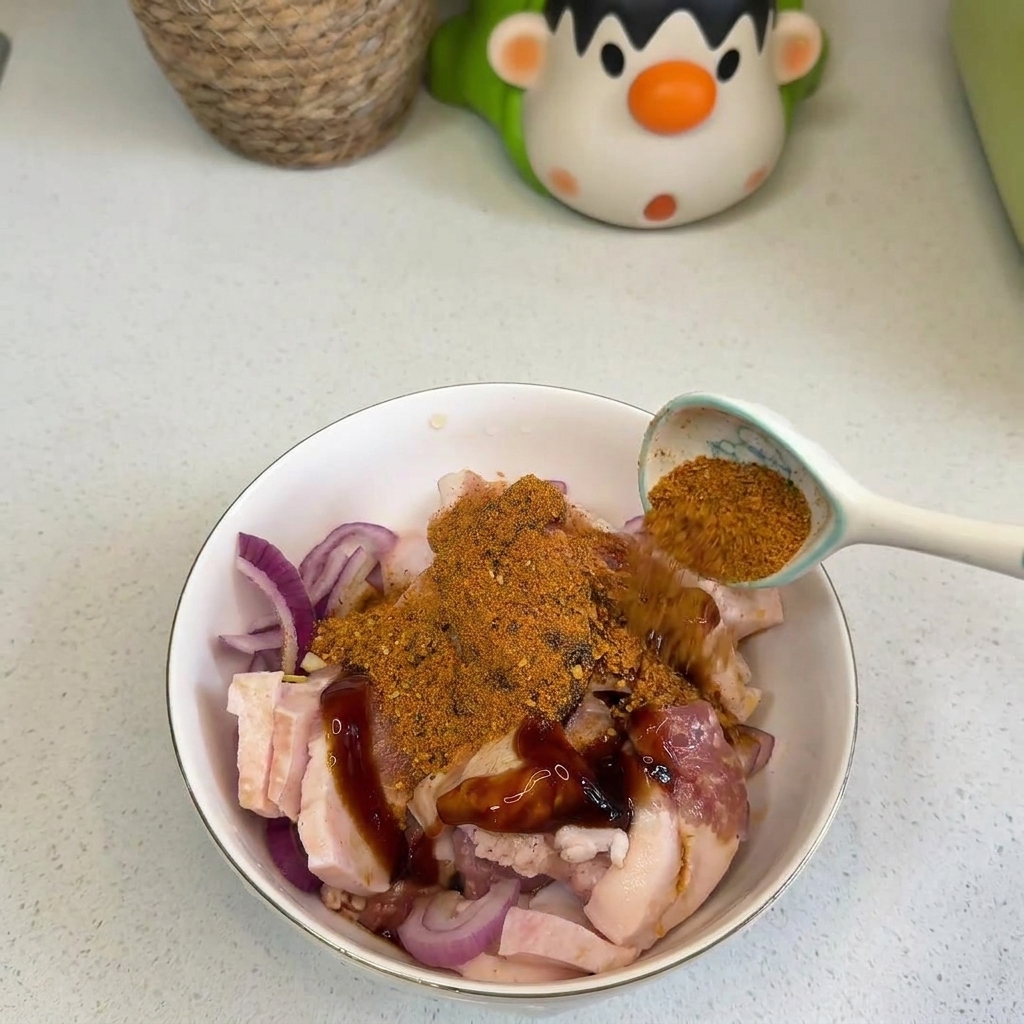 Eine Hand streut mit einem Löffel orangefarbenes BBQ-Gewürz und rotes Chilipulver über rohe Schweinebauchscheiben und rote Zwiebeln in einer weißen Schüssel.