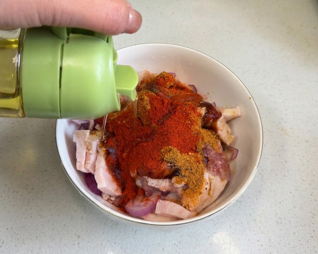 Helles Speiseöl wird aus einem Behälter mit grünem Deckel über den mit roten Gewürzen bestreuten Schweinebauch geträufelt.