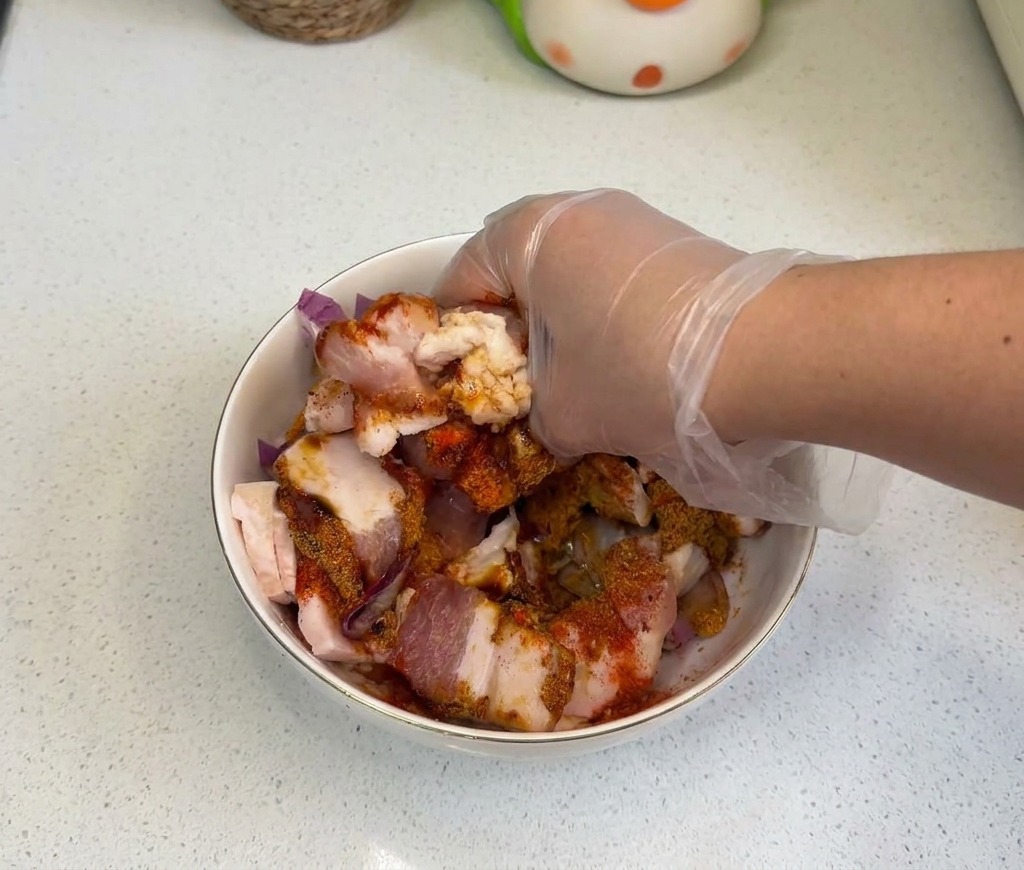 Eine behandschuhte Hand vermengt rohen Schweinebauch, Zwiebeln und Gewürze gründlich in einer weißen Schüssel.
