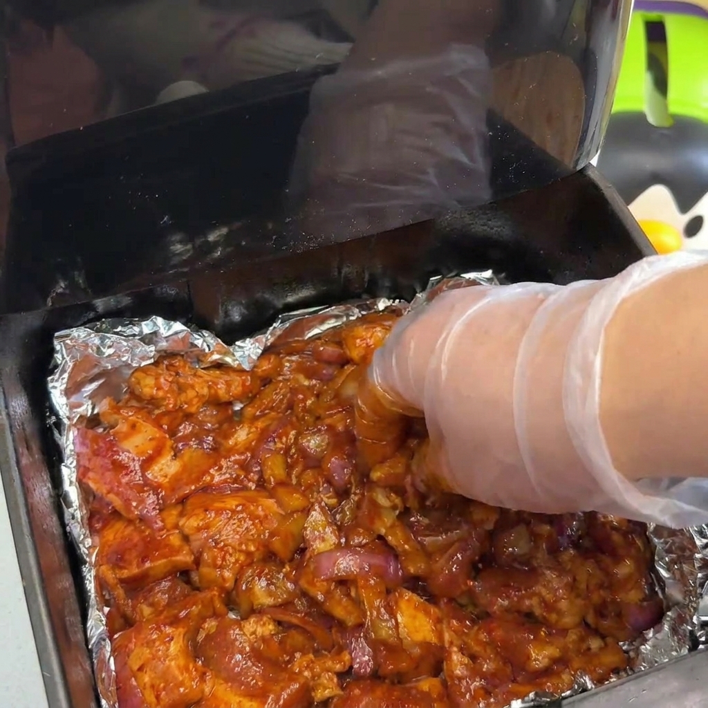 Eine behandschuhte Hand verteilt eine dicke Schicht aus mariniertem Schweinefleisch und Zwiebeln im Korb einer Heißluftfritteuse.