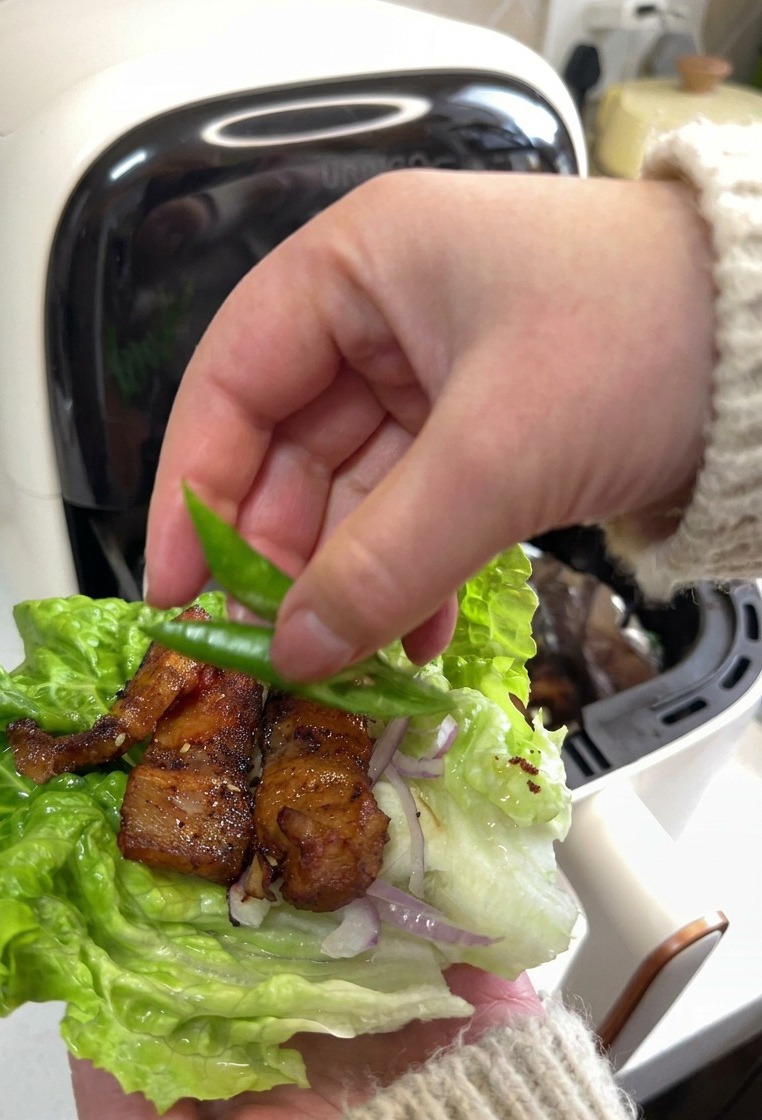 Eine Hand legt zwei leuchtend grüne Chilischeiben auf ein Salatblatt, das mit geröstetem Schweinebauch und in Scheiben geschnittenen roten Zwiebeln gefüllt ist.