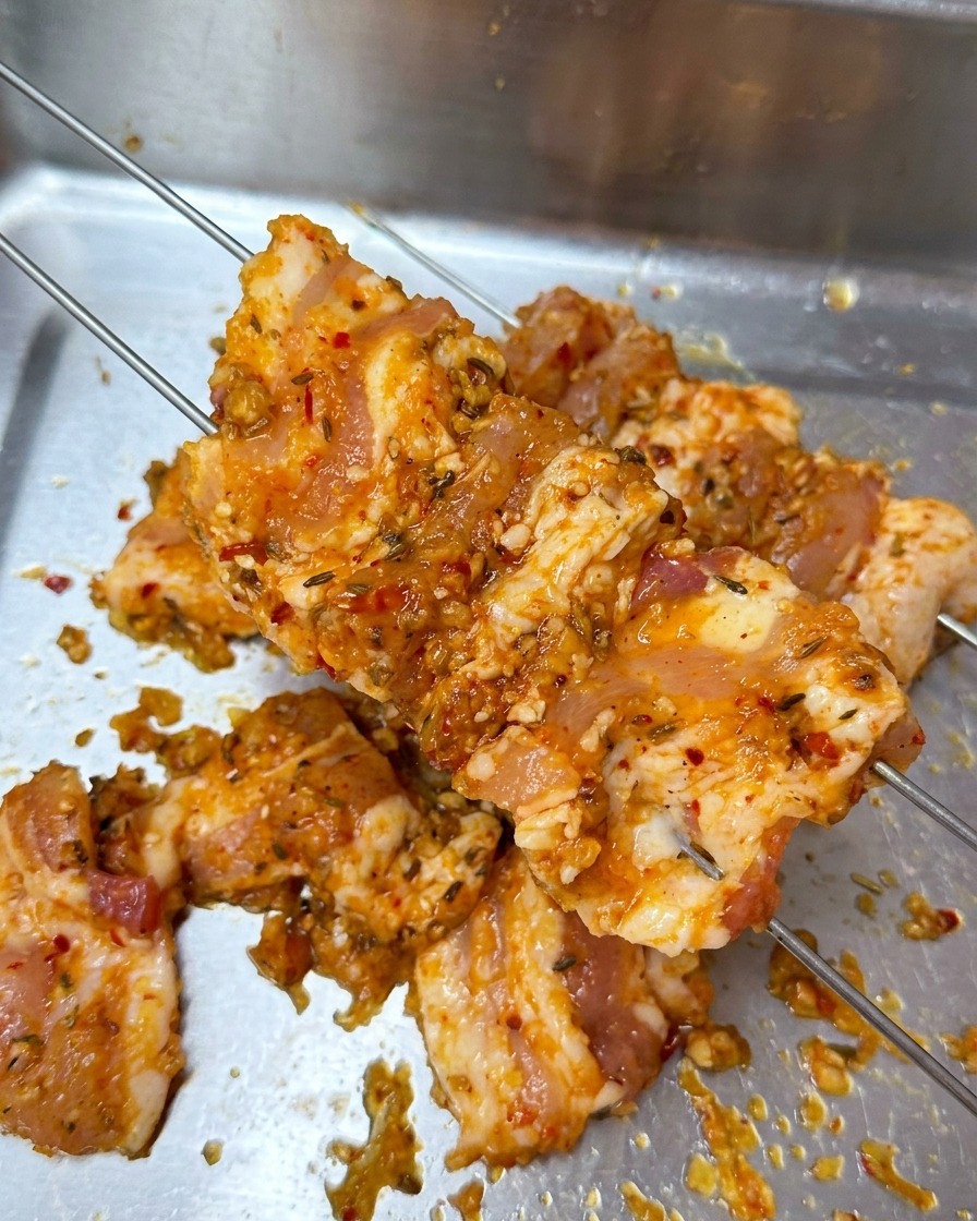 Marinierte Schweinebauchscheiben werden auf zwei dünne Metallspieße über einem Tablett gesteckt.
