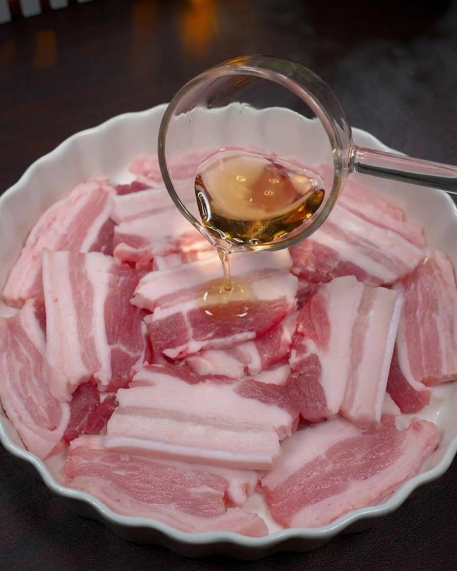 Klare Flüssigkeit wird aus einer Schöpfkelle über rohe Schweinebauchscheiben gegossen.