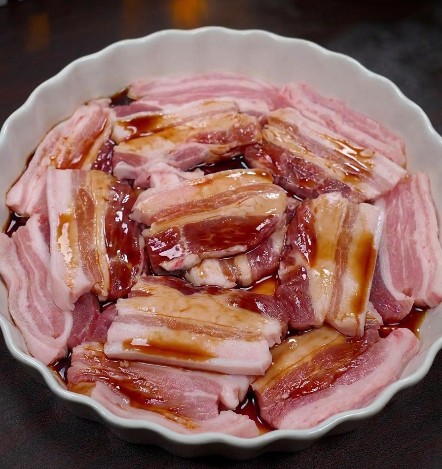 Schweinebauchscheiben in einer weißen Schale werden mit einer dunklen Marinade überzogen.