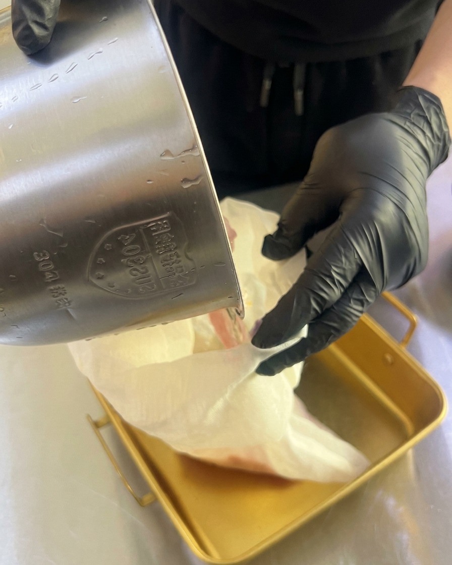Hände in Handschuhen drücken ein weißes Tuch aus, um frischen Fruchtsaft in eine goldene Metallschale zu filtern.