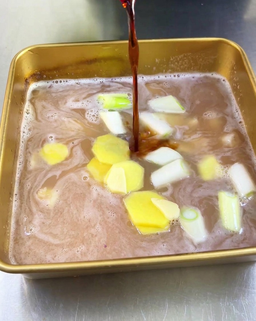 Dunkle Sojasauce wird in eine goldene Schale gegossen, die bereits hellen Fruchtsaft, Ingwerscheiben und Frühlingszwiebeln enthält.