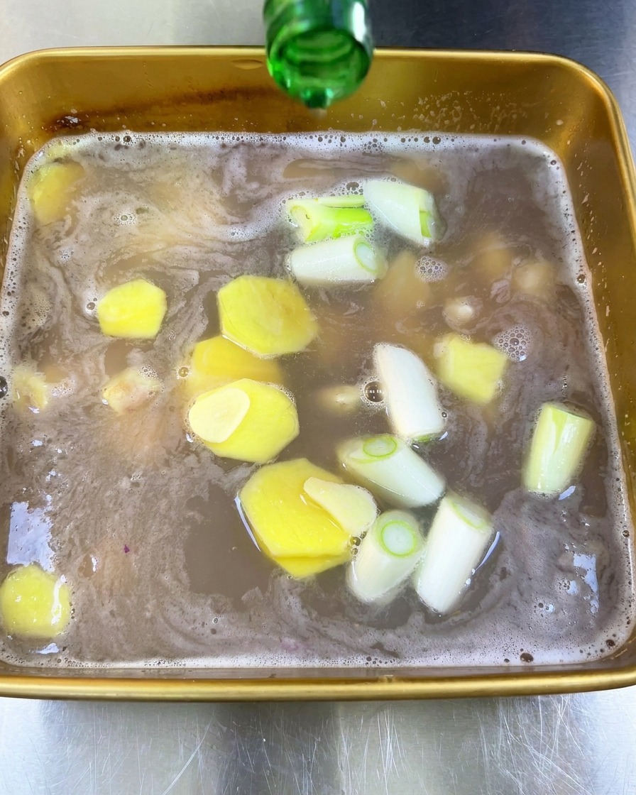Klarer Reiswein wird aus einer grünen Glasflasche in die Marinade mit Ingwer und Schalotten gegossen.