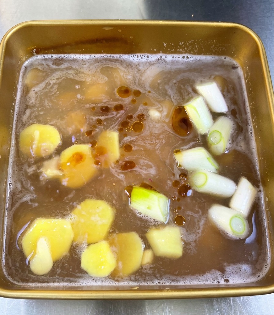 Draufsicht auf eine goldbraune Sojamarinade mit Ingwerscheiben, Frühlingszwiebeln und dunklen Sesamöltropfen auf der Oberfläche.