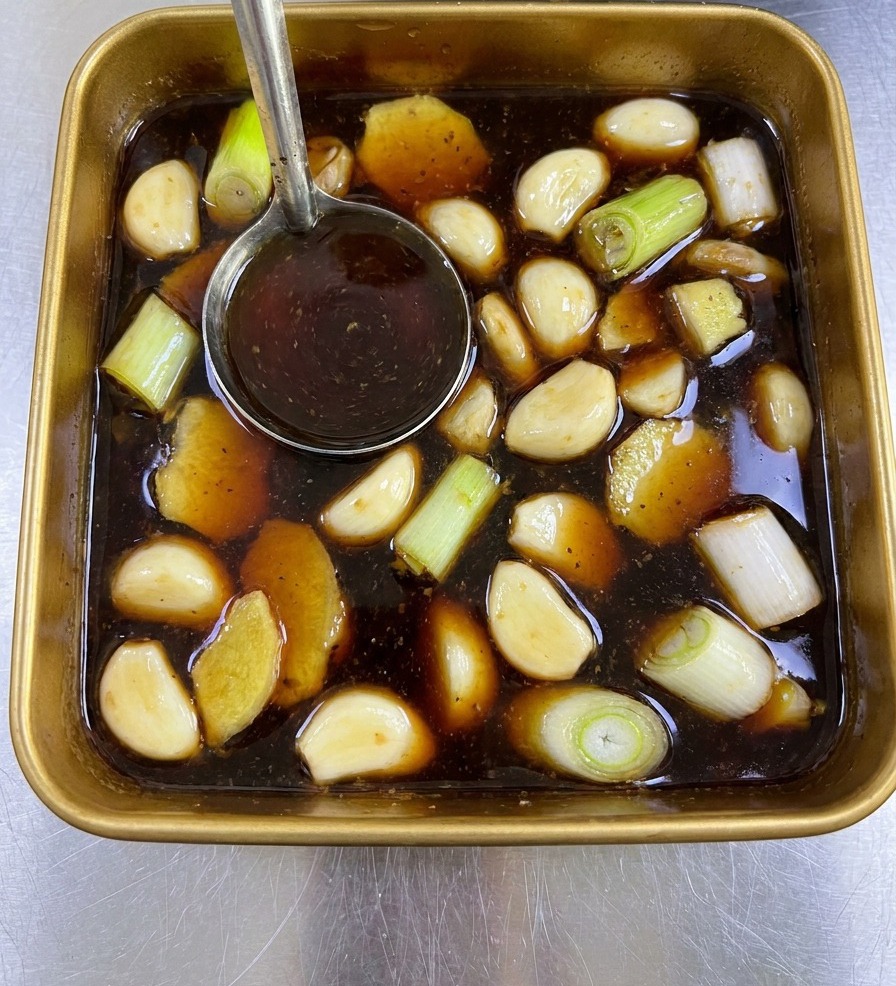 Eine Metallkelle rührt die dunkelbraune Marinade mit Ingwer und Frühlingszwiebeln in einer quadratischen goldenen Schale um.