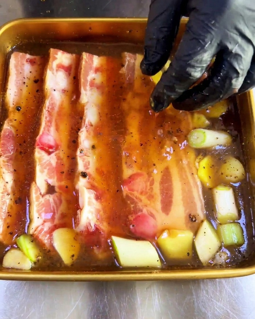Eine Hand in einem Handschuh drückt dicke, rohe Schweinebauchstreifen in die dunkle Marinade mit Aromaten.