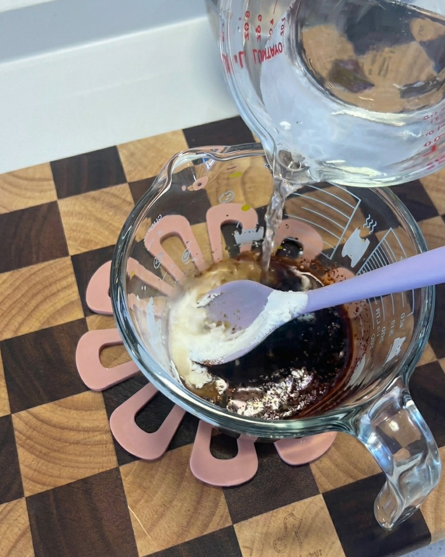 Wasser wird aus einem Glasbehälter in eine Tasse mit einer dunklen Mischung aus Sojasauce und Gewürzen gegossen.