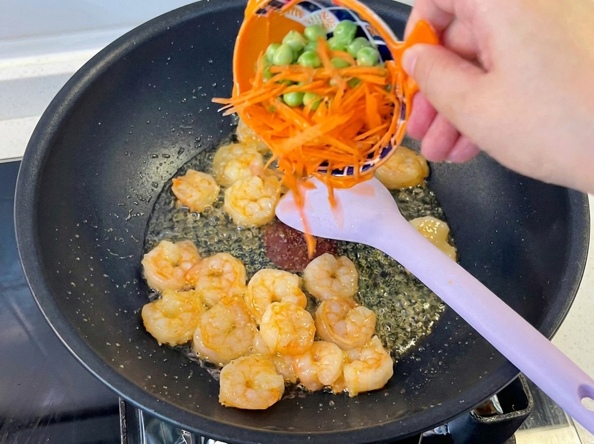 Eine Hand gibt eine Mischung aus Karottenstiften und Erbsen in den Wok zu den gebratenen Garnelen.