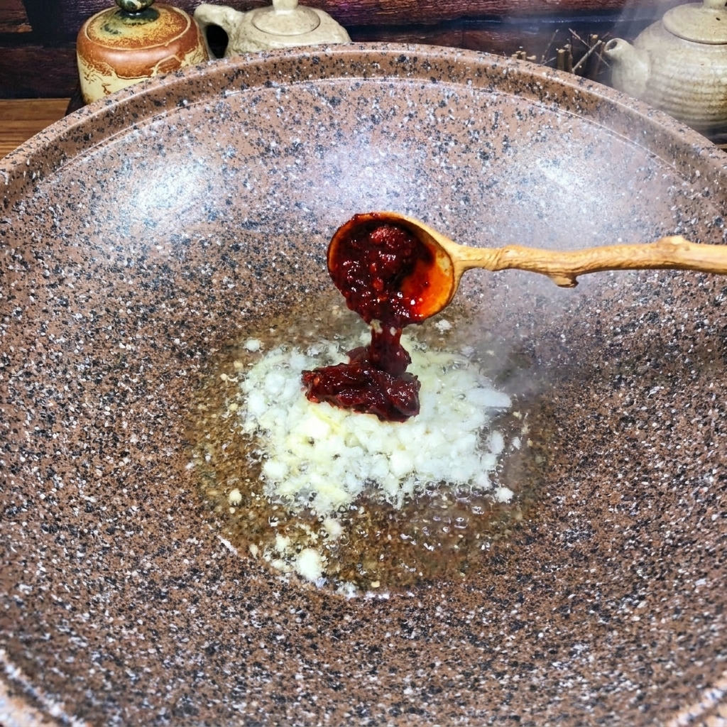 Ein Holzlöffel gibt rote Chili-Bohnenpaste in einen Wok zu den angebratenen Aromaten.