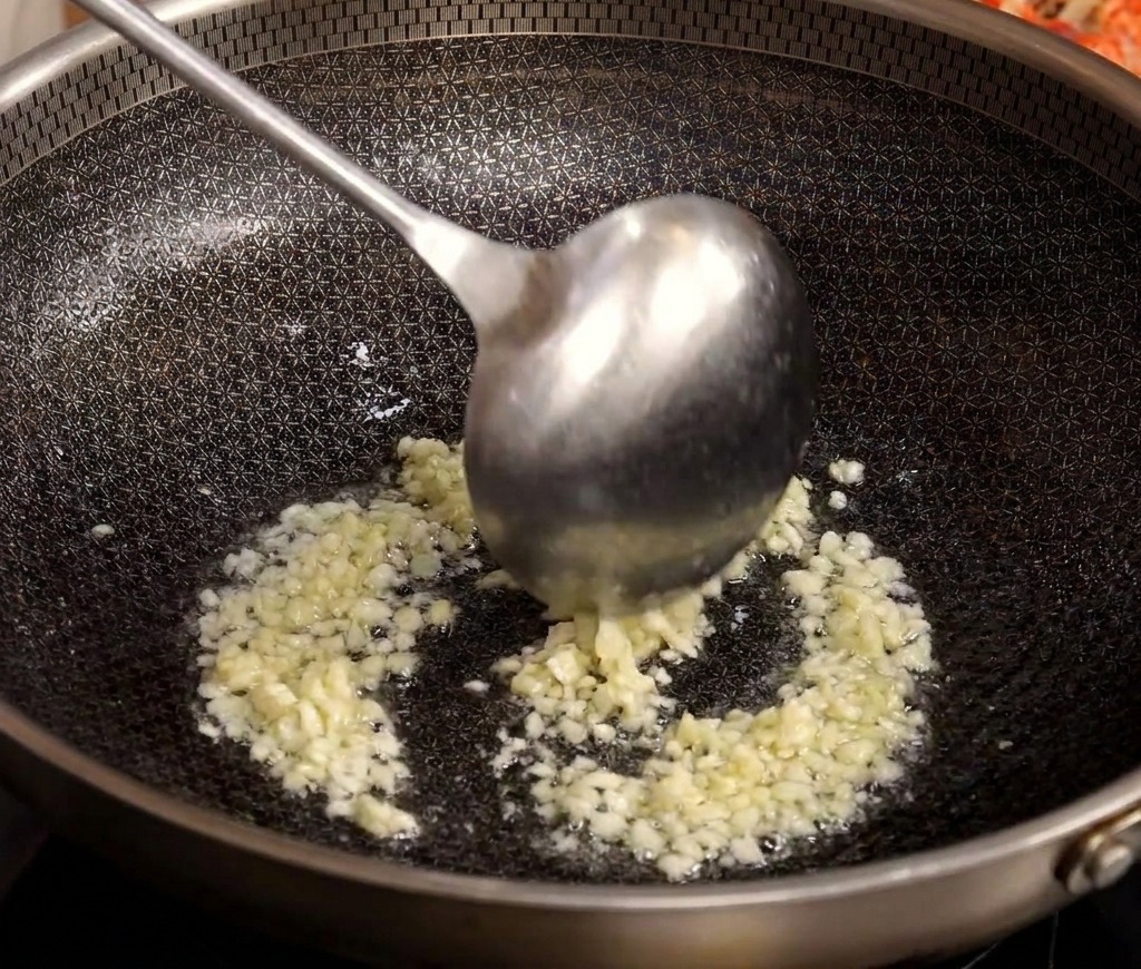 Ein Metall-Löffel rührt gehackten Knoblauch in einem heißen Wok mit Öl um.