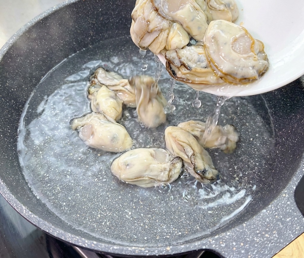 Rohe, pralle Austern gleiten von einem weißen Teller in einen Wok mit kaltem Wasser.
