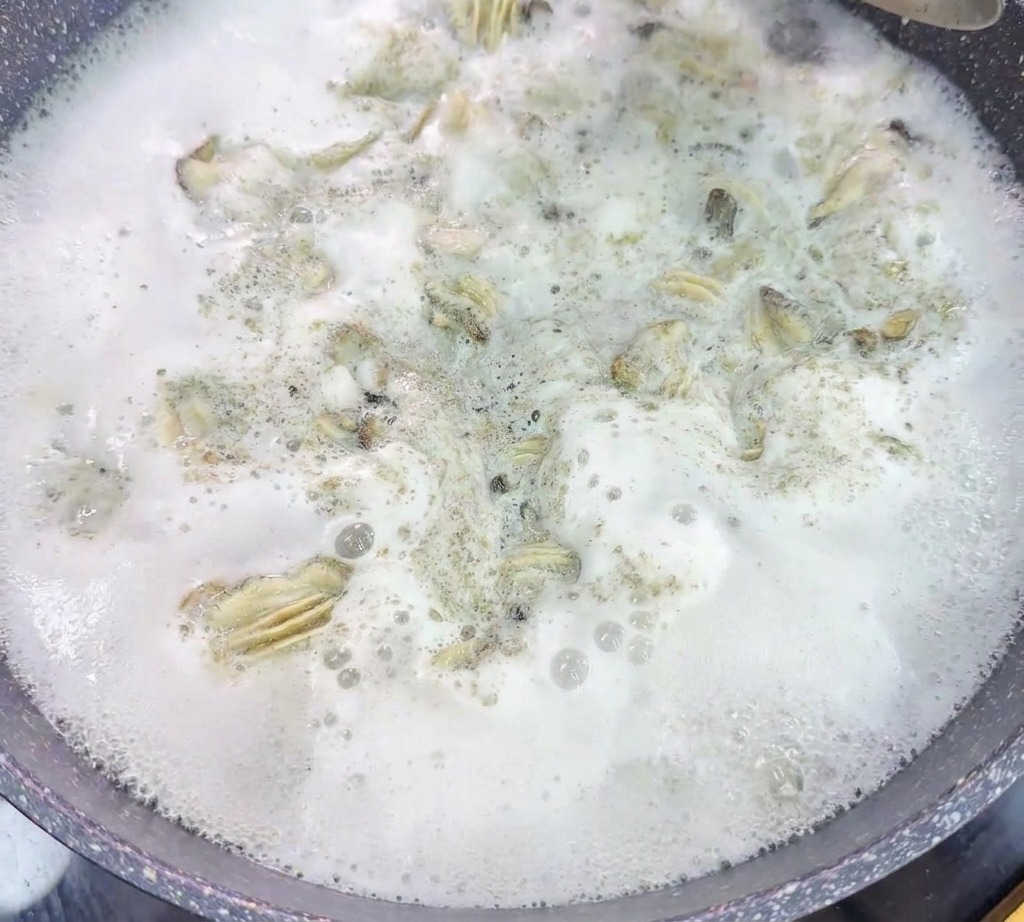 Ein Metallsieb hebt blanchierte Austern aus einem Topf mit kräftig kochendem, schaumigem Wasser.