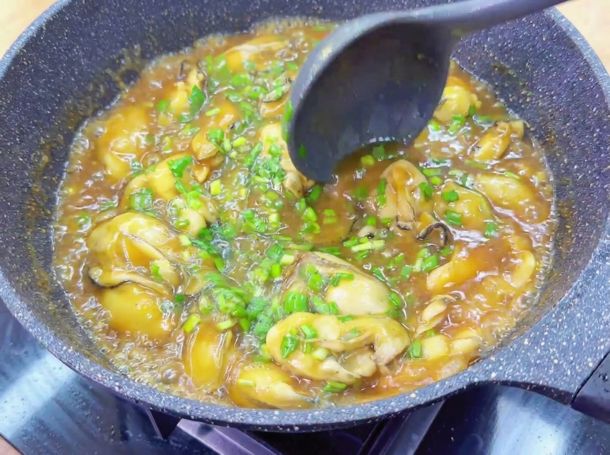 Eine dunkle Kelle rührt Austern und Schnittknoblauch in einer glänzenden braunen Sauce um.