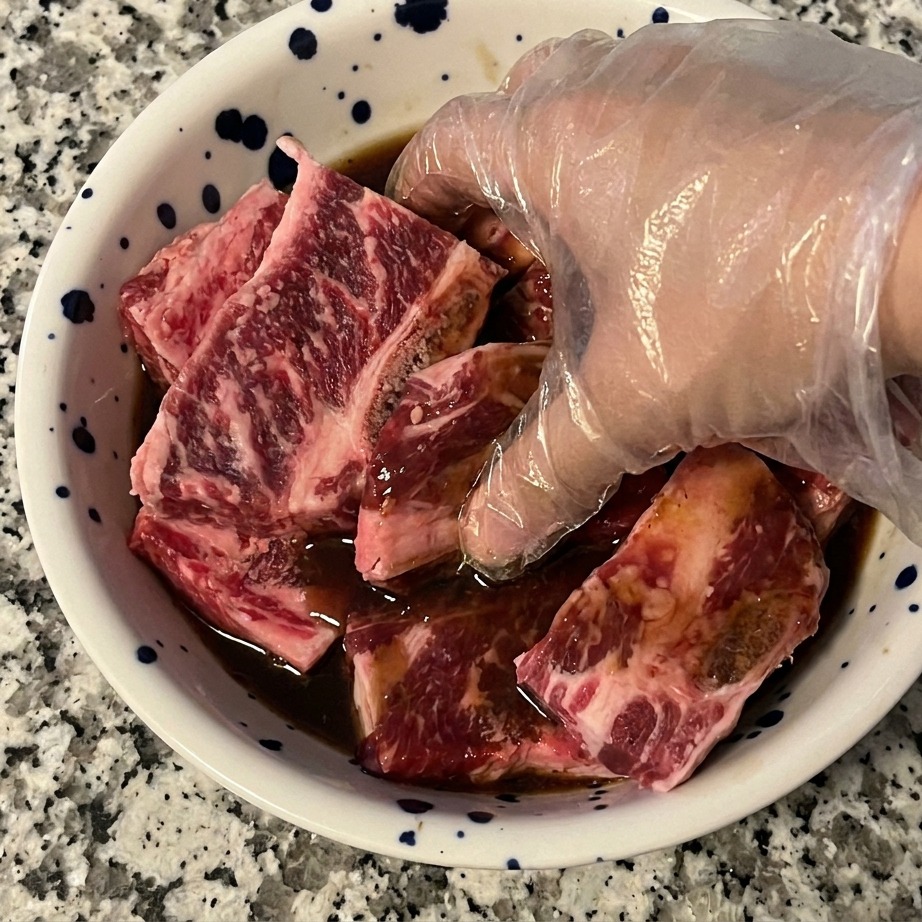 Eine Hand mit Handschuh vermengt die Rinderrippen gründlich mit der dunklen Marinade in einer Schüssel.