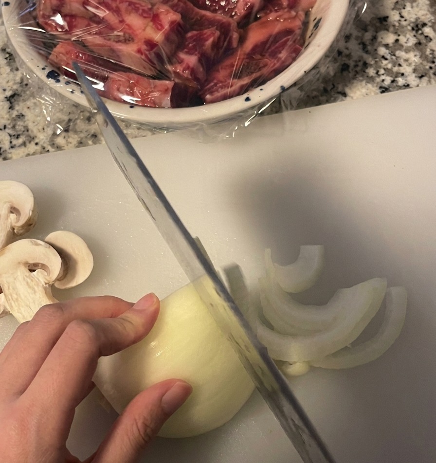 Eine weiße Zwiebel wird mit einem Messer auf einem Brett in feine Halbmonde geschnitten.