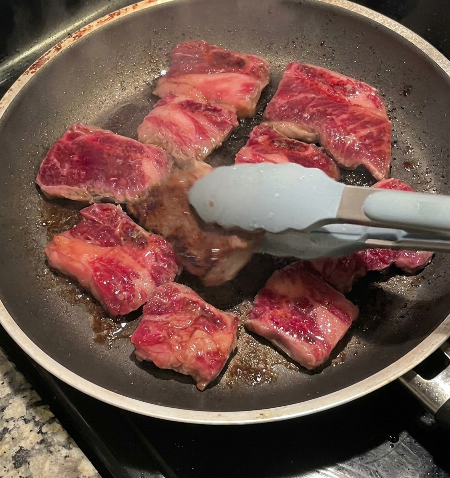 Zangen wenden die Rippchen in der Pfanne und zeigen die dunkelbraune, karamellisierte Kruste.
