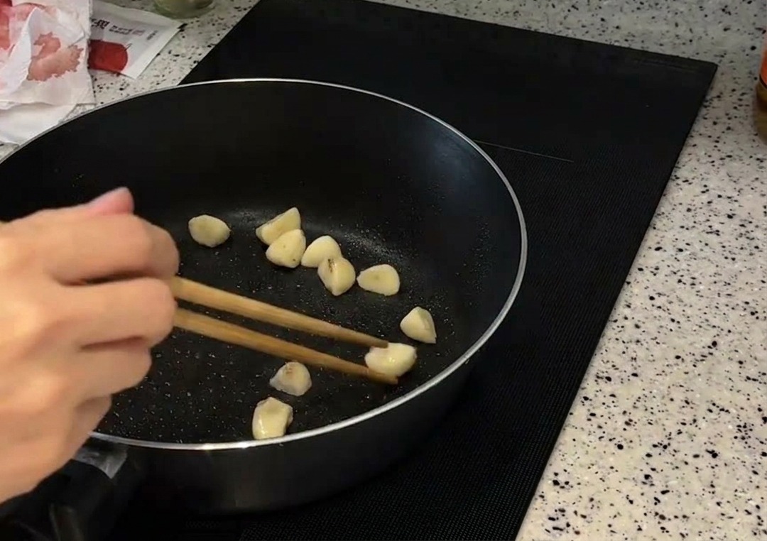 Ganze Knoblauchzehen werden mit Stäbchen in einer dunklen Pfanne goldbraun gebraten.