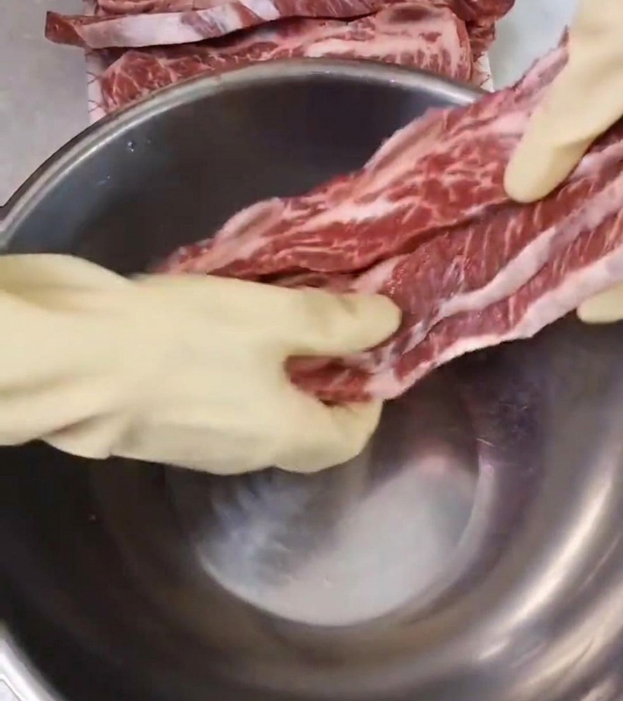 Eine Person mit weißen Handschuhen legt rohe, marmorierte Rinderrippen in eine tiefe Edelstahlschüssel auf einer sauberen Arbeitsplatte.