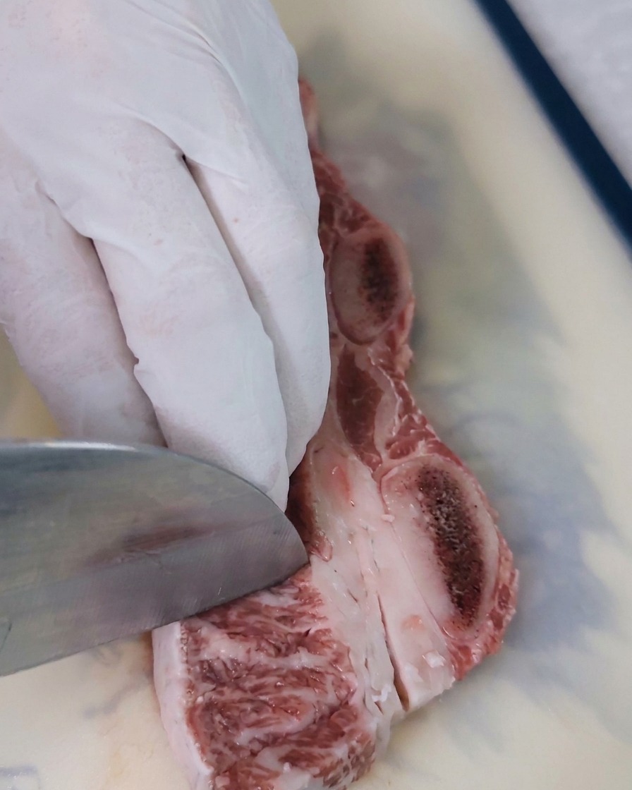 Nahaufnahme eines Messers, das diagonale Schnitte in das rote Fleisch einer Rinderrippe macht, um die Oberfläche für die Marinade vorzubereiten.