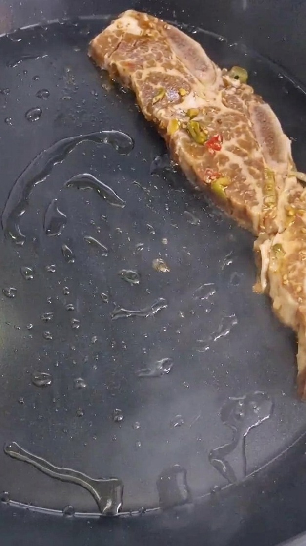 Eine marinierte Rinderrippe mit Paprika- und Zwiebelstücken wird in eine heiße, geölte Pfanne gelegt.