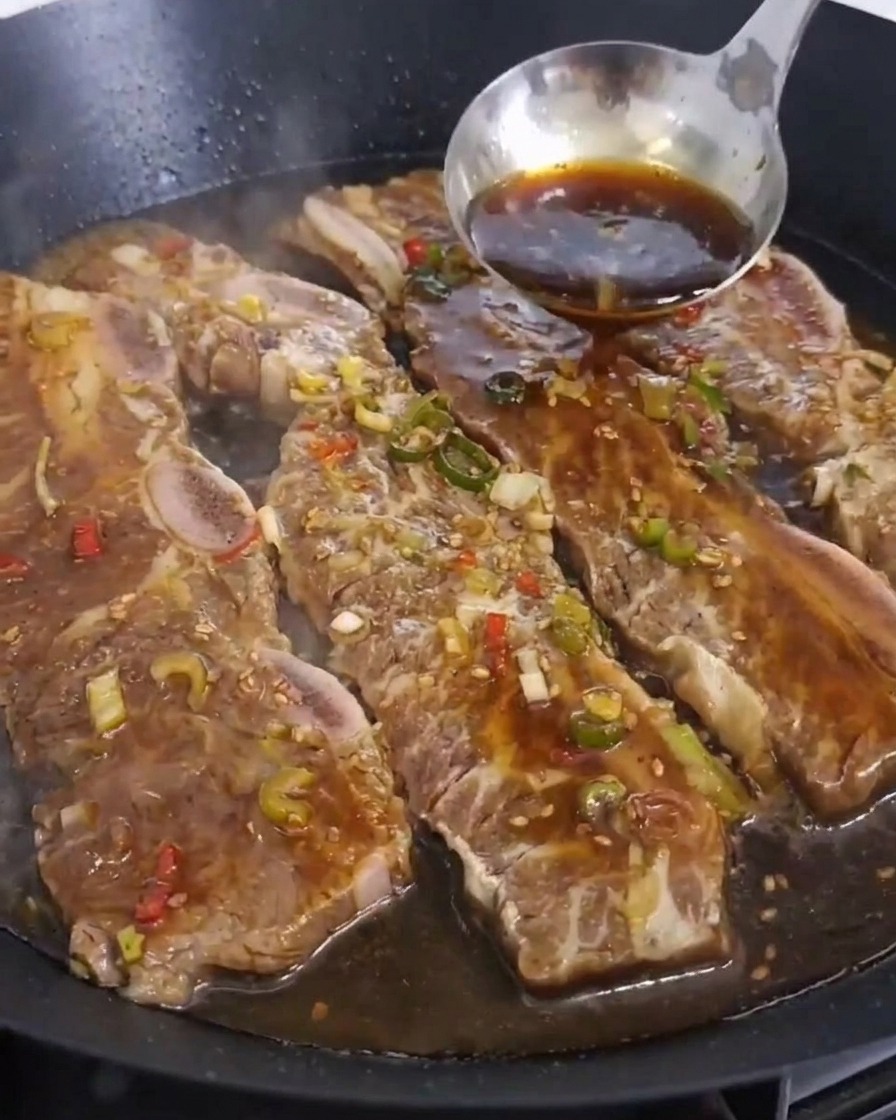Eine Schöpfkelle aus Edelstahl gießt dunkle Marinade über mehrere Rinderrippen, die in einer Pfanne garen.