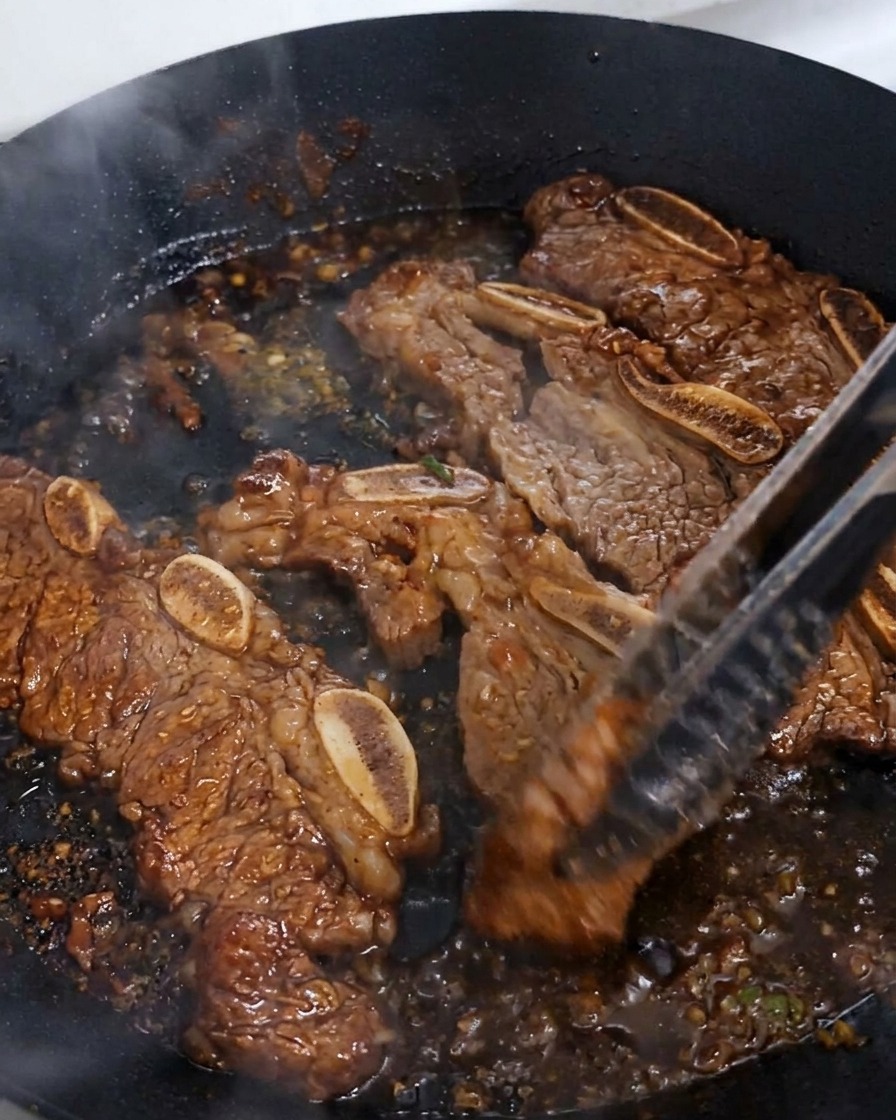 Rinderrippen werden mit einer Zange in einer Pfanne gewendet, während die dunkle Sauce einkocht und eine glänzende Glasur bildet.