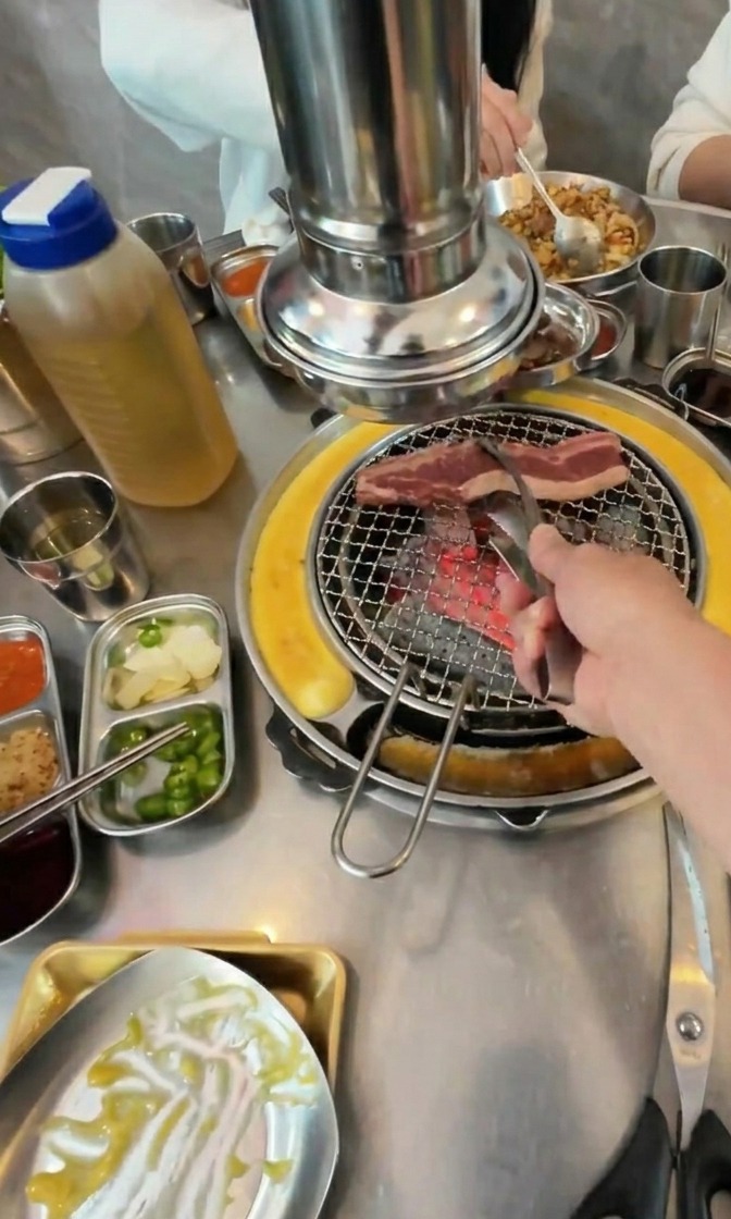 Eine Hand legt mit einer Metallzange rohe, fettige Rindfleischstreifen auf einen heißen Holzkohlegrill.