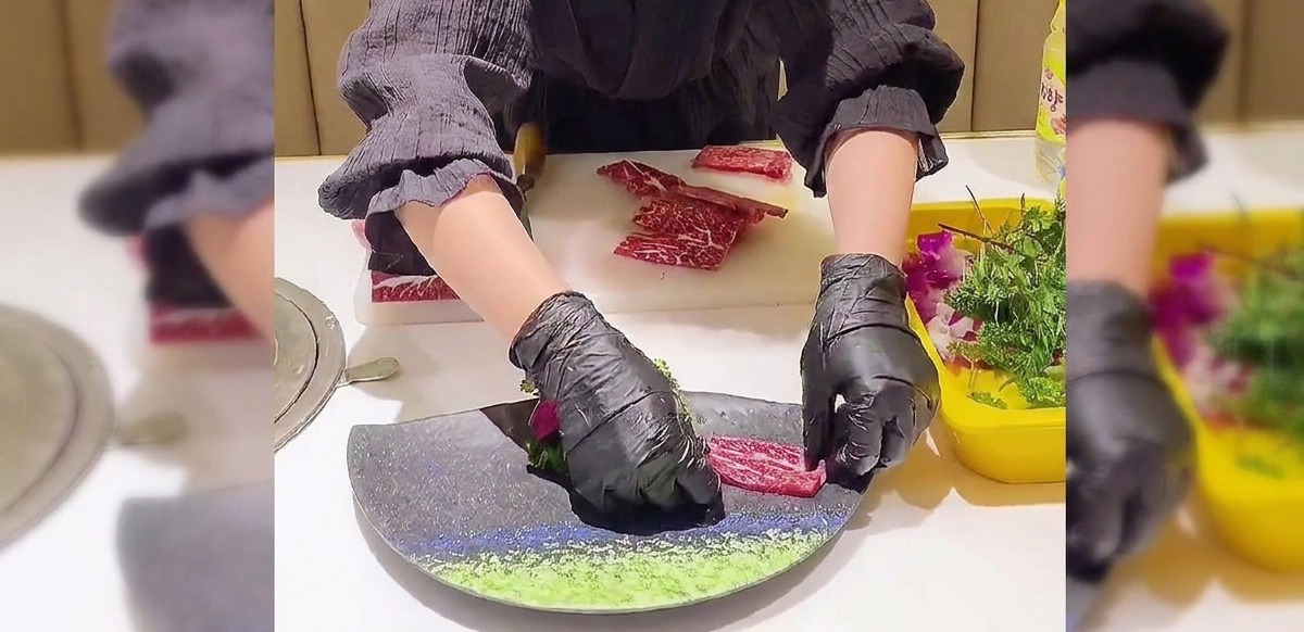 Hände in schwarzen Handschuhen schichten dünne, marmorierte Rindfleischscheiben überlappend in Fächerform auf einem dunklen Teller.