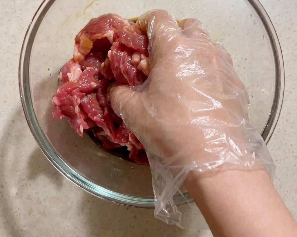Eine Hand mit einem Einweghandschuh massiert die Marinade in die rohen Schweinefleischstreifen ein.