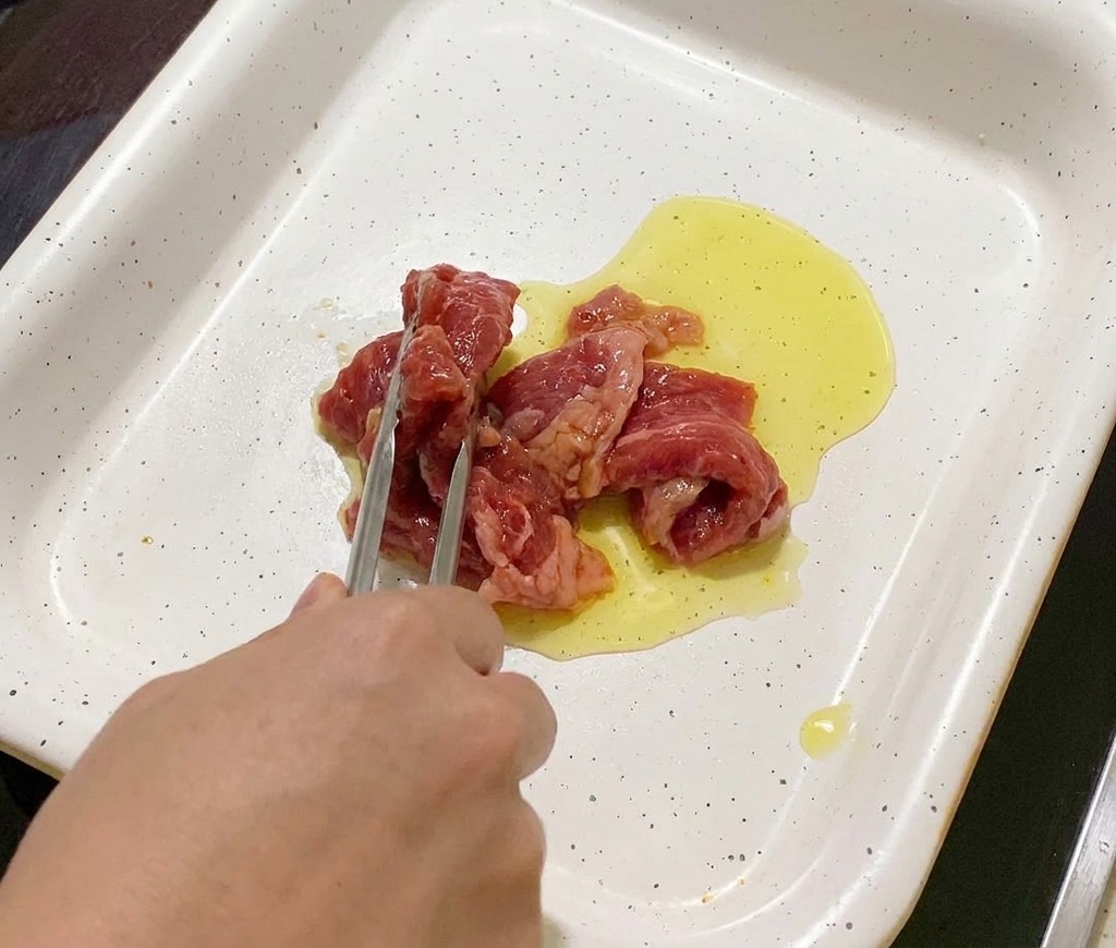 Fleischstreifen werden mit einer Grillzange in eine heiße Pfanne mit glänzendem Öl gelegt.
