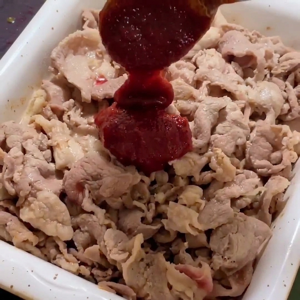 Die dicke rote Chilisauce wird über das angebratene Fleisch in der weißen Pfanne gegeben.