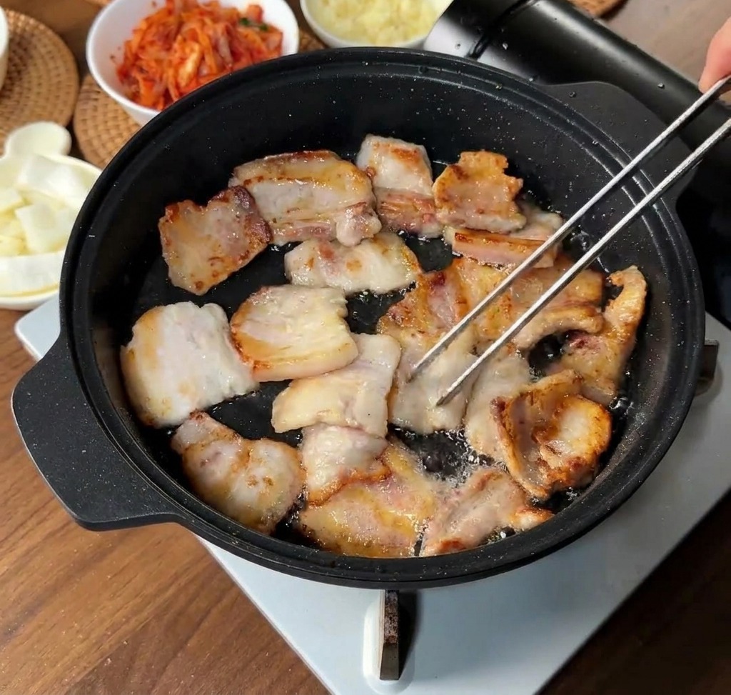 Schweinebauchscheiben bräunen im eigenen Fett in einer schwarzen Pfanne; einige Stücke werden mit einer Zange gewendet.