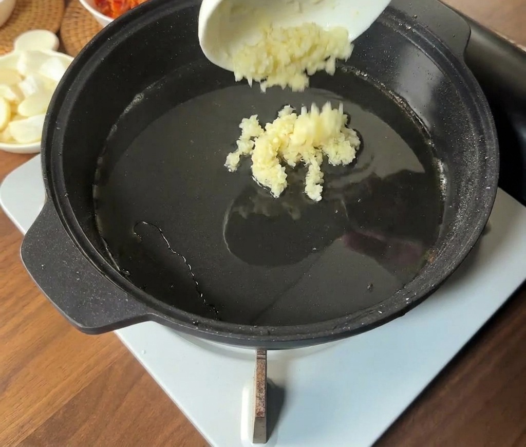 Gehackter Knoblauch wird aus einer kleinen Schüssel in die heiße, fettige Mitte der Pfanne gegeben.