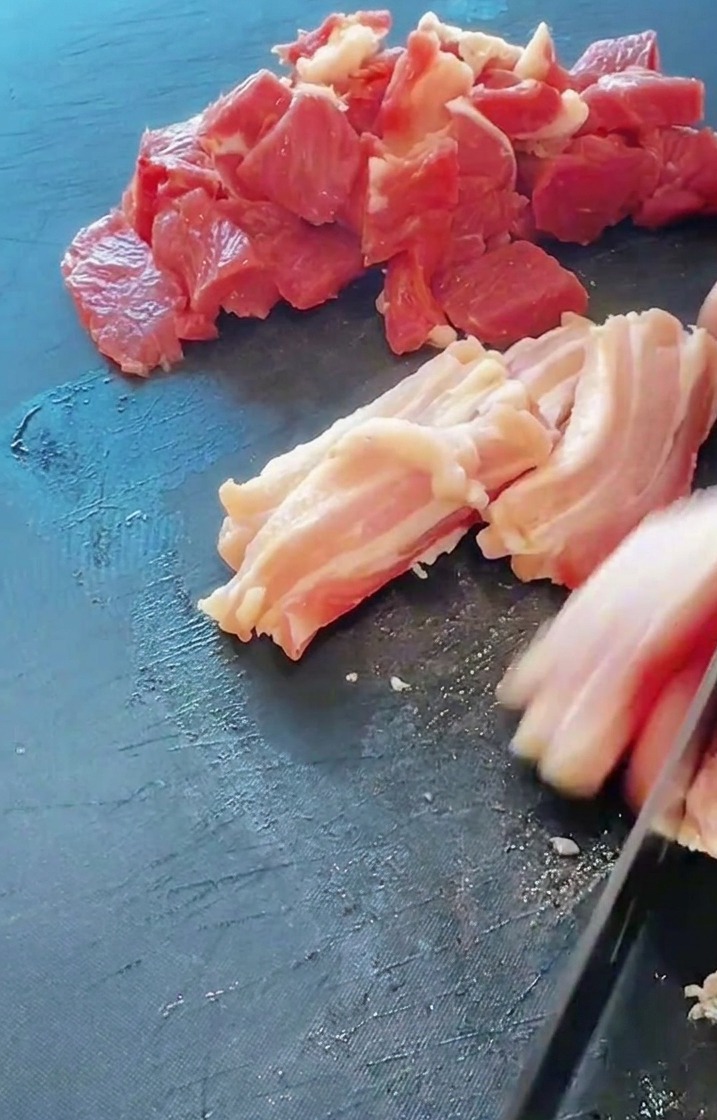 Nahaufnahme eines Messers, das frischen Schweinebauch auf einem dunklen Schneidebrett in Streifen schneidet.