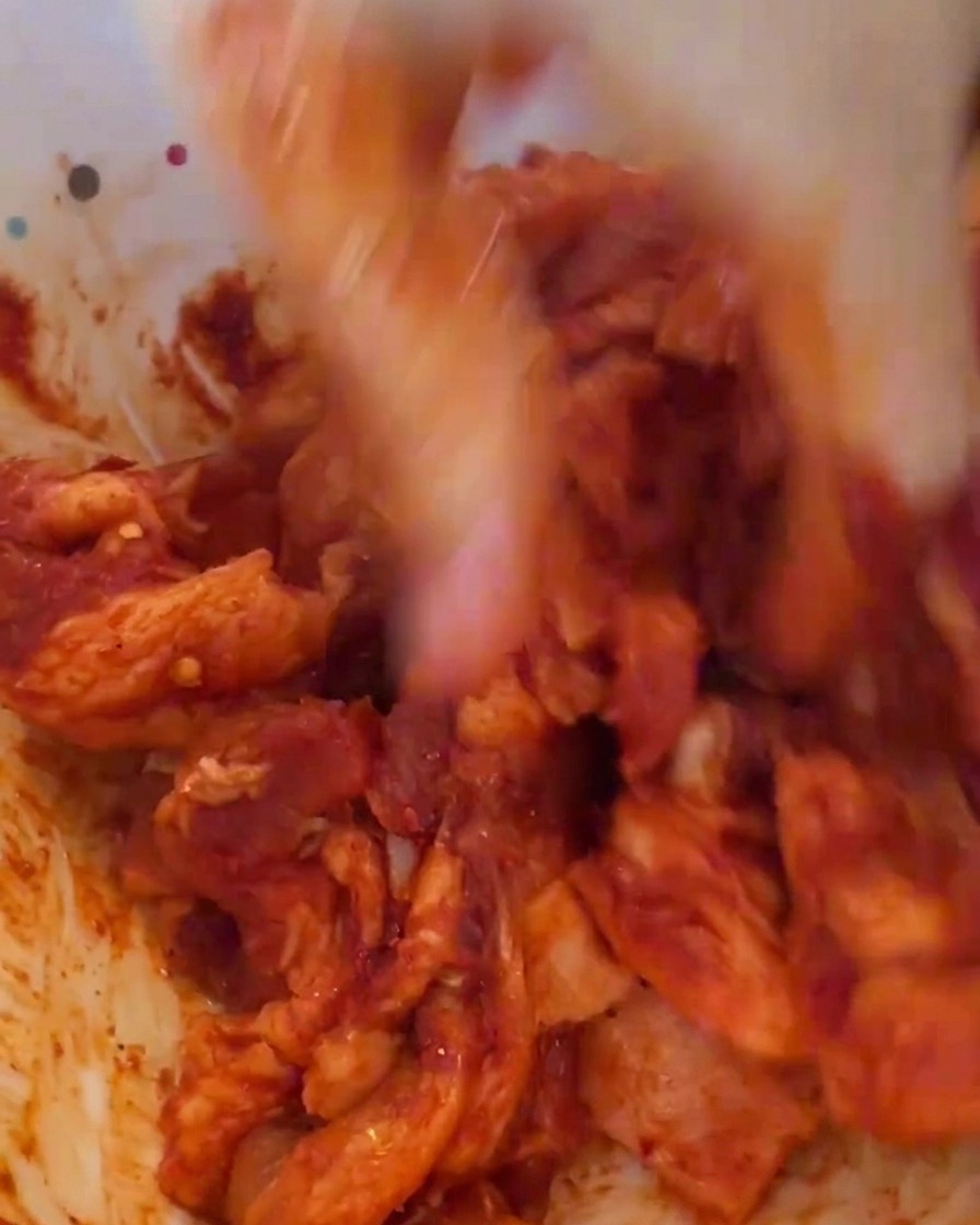 Eine Hand mit Handschuh vermengt das rote marinierte Fleisch in einer weißen Schüssel.