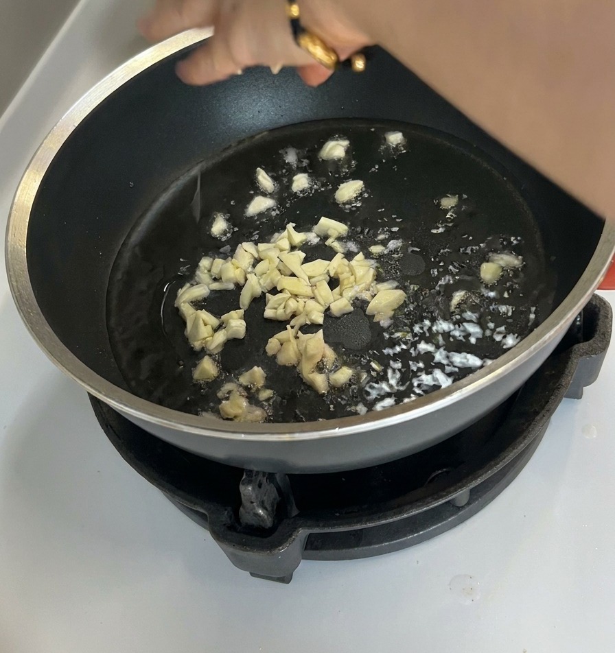 Gehackter Knoblauch brät in Öl am Boden einer dunklen, beschichteten Pfanne.