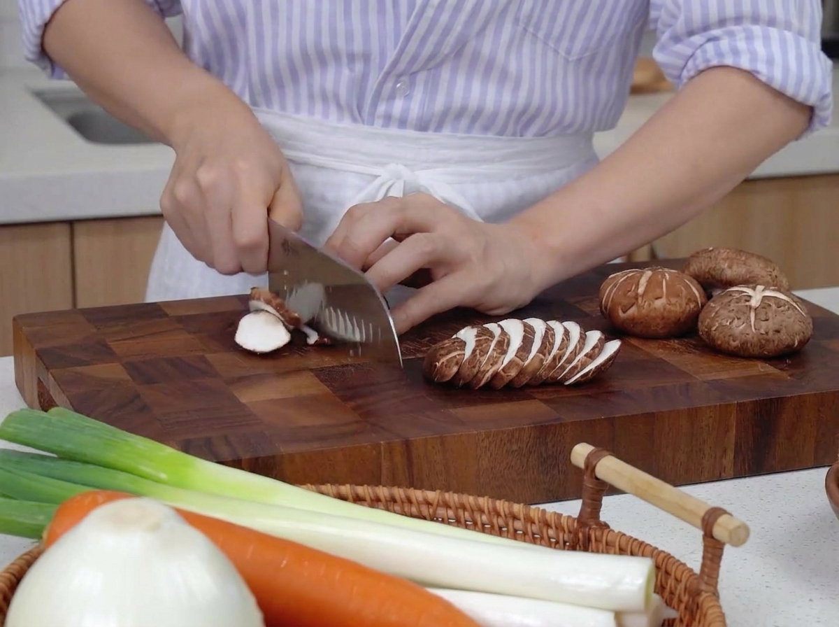Hände schneiden Shiitake-Pilze auf einem dunklen Holzbrett neben Karotten und Frühlingszwiebeln.